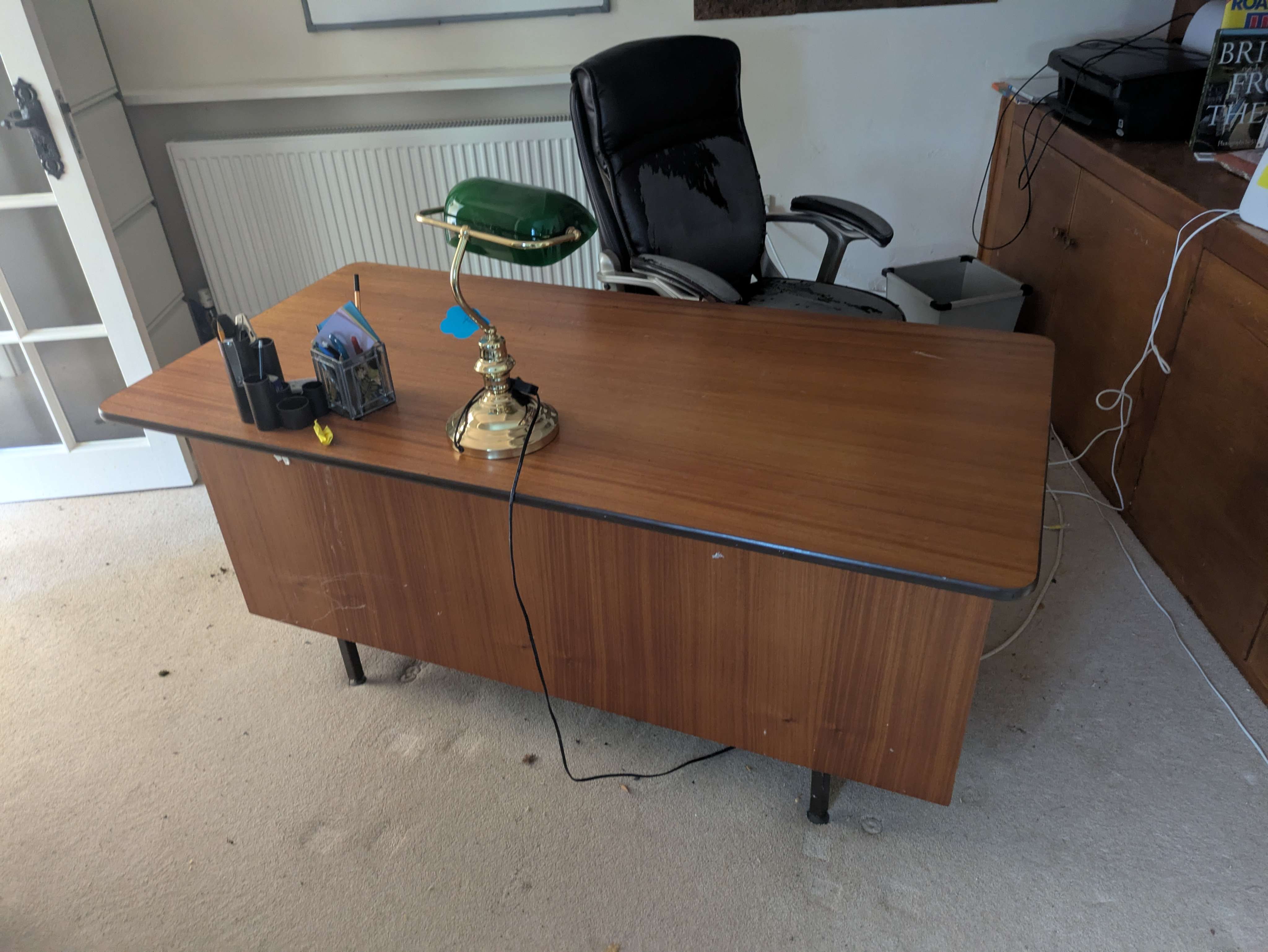 A vintage teak office desk and brass-and-green banker's lamp left bare in a room mid-clearance