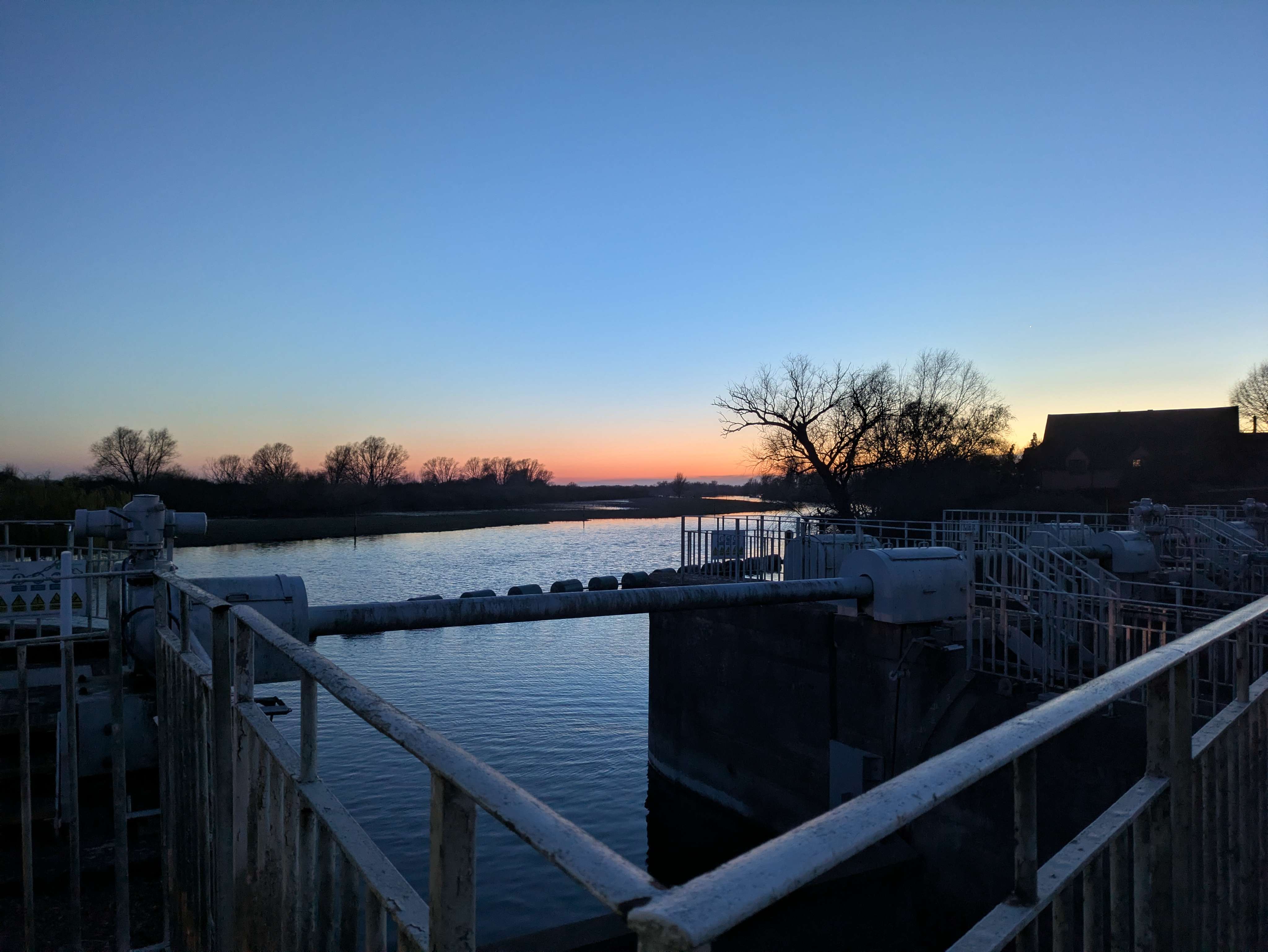 Pastel sunset over a Cambridgeshire sluice and river — silhouettes of trees, water glassy, work done