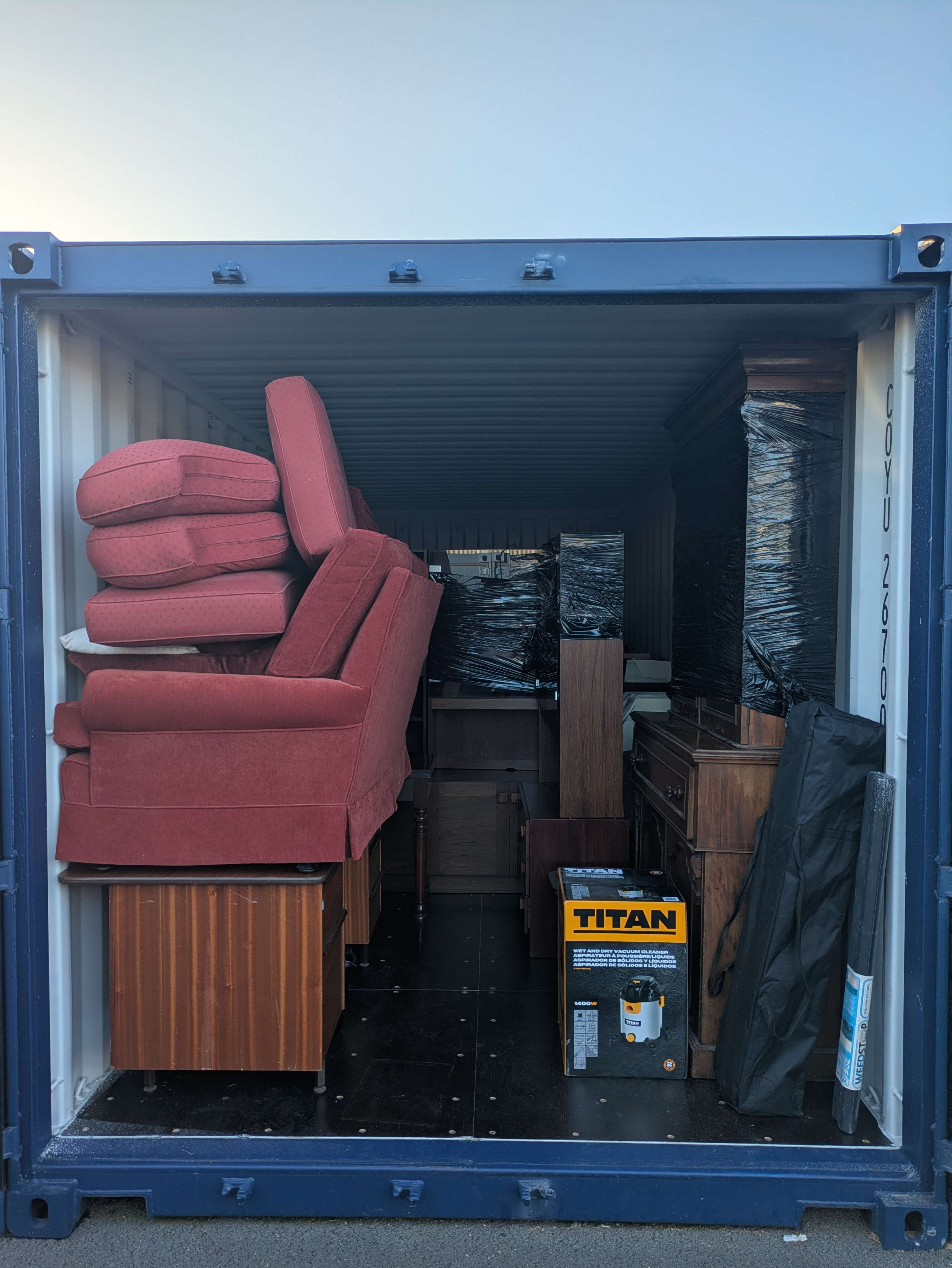 A blue storage container packed with stacked armchair cushions, a wrapped wardrobe, a sideboard and a boxed Titan vacuum cleaner