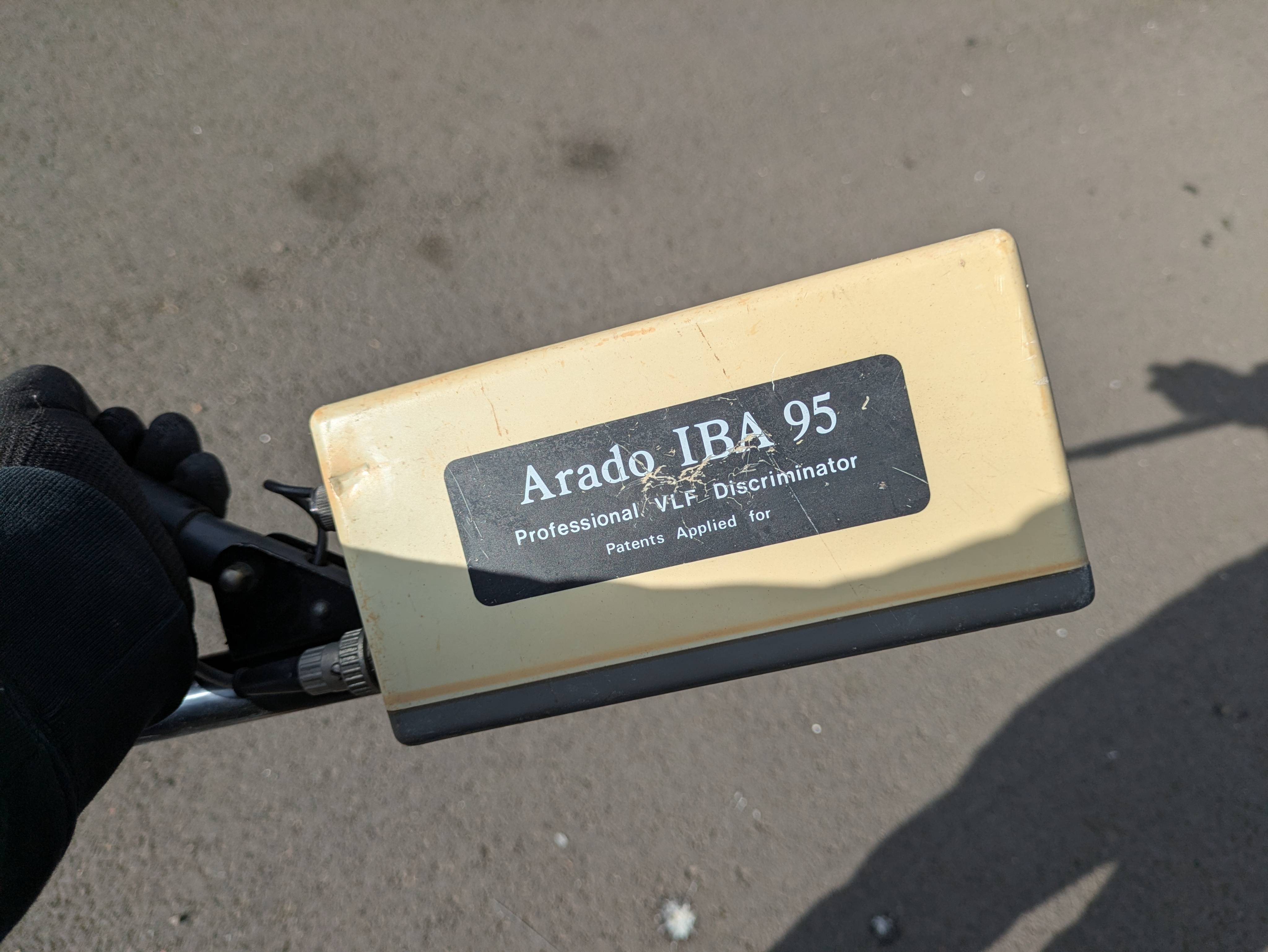 A gloved hand holding a vintage Arado IBA 95 metal detector control box, scratched yellow body, dark tarmac below